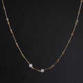 A delicate gold chain necklace featuring three small, round, pale blue-gray beads spaced along the front, displayed against a dark gray background.