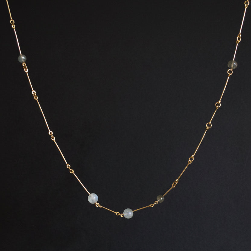 A delicate gold chain necklace featuring three small, round, pale blue-gray beads spaced along the front, displayed against a dark gray background.