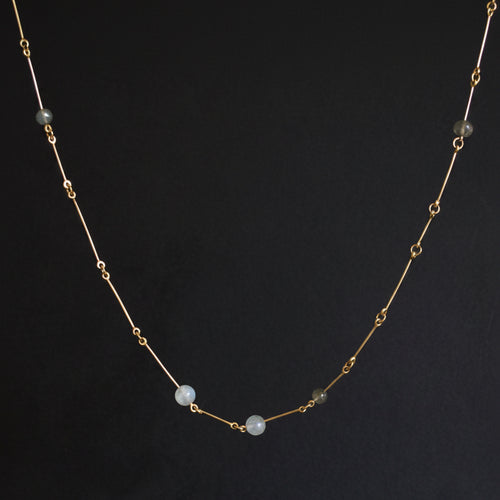A delicate gold chain necklace featuring three small, round, pale blue-gray beads spaced along the front, displayed against a dark gray background.