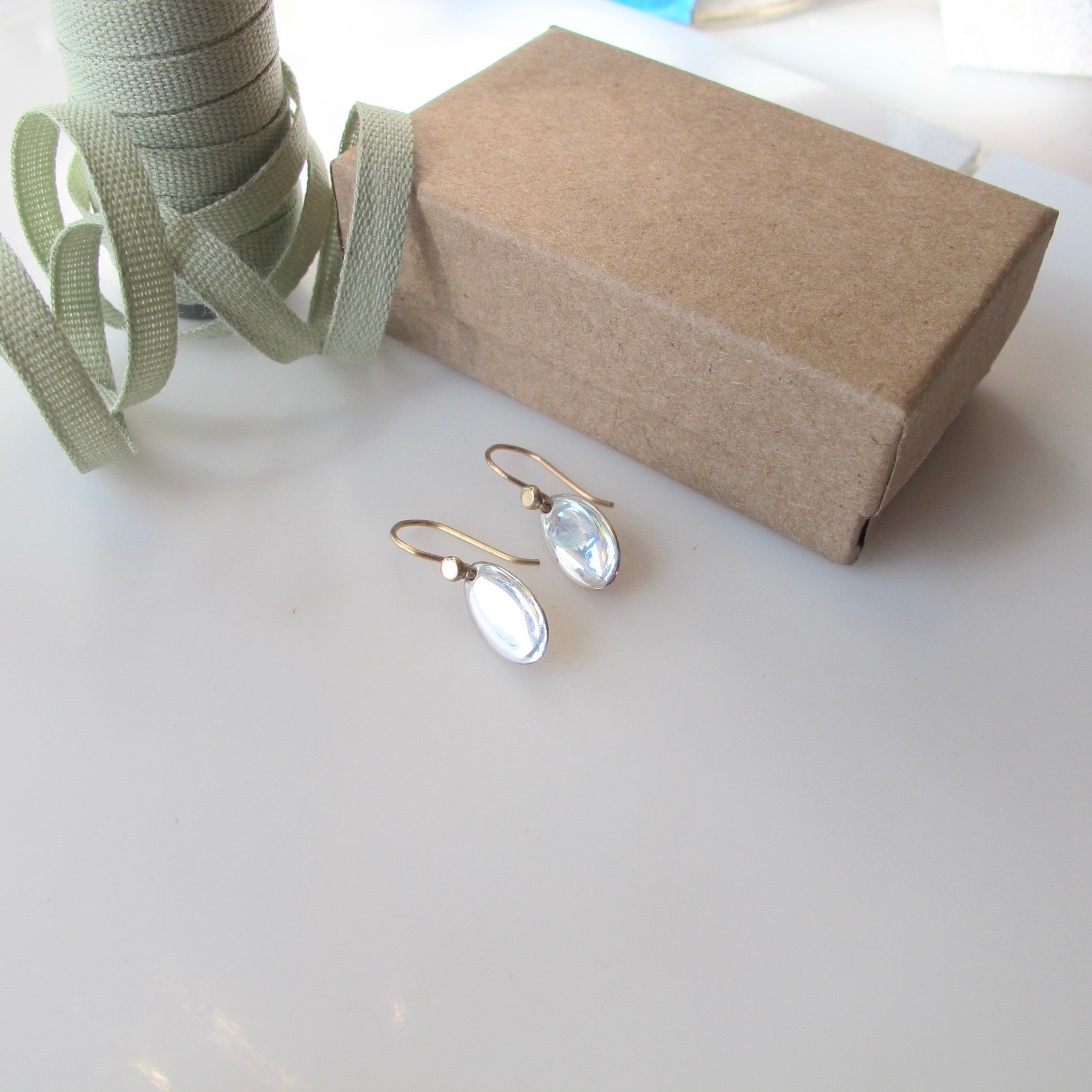 A pair of gold-tone drop earrings with oval clear stones, displayed on a white surface next to a brown kraft gift box and a spool of sage green ribbon.