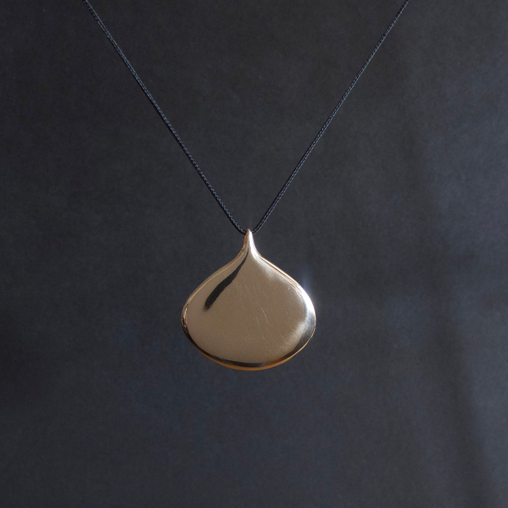 A minimalist teardrop-shaped pendant necklace in polished gold, suspended from a thin dark cord against a dark gray background.