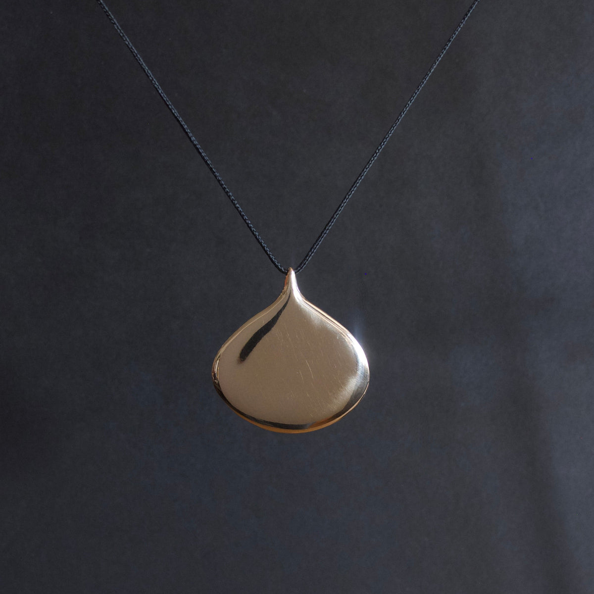A minimalist teardrop-shaped pendant necklace in polished gold, suspended from a thin dark cord against a dark gray background.