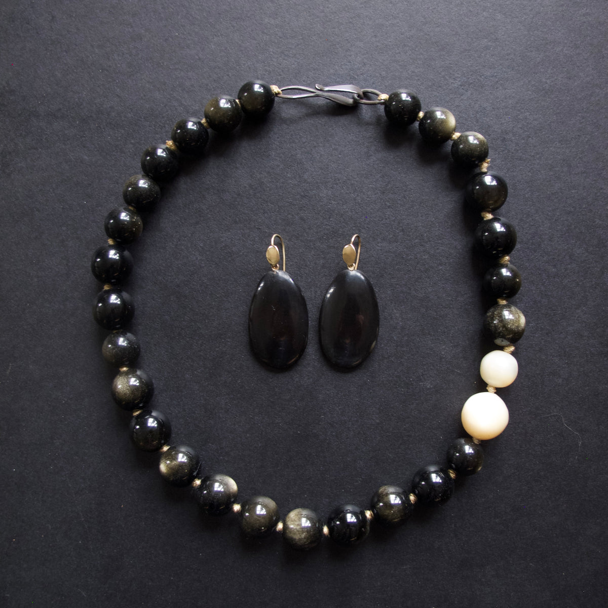 A beaded necklace and matching drop earrings laid flat on a dark textured background. The necklace and earrings use glossy black and gray marbled beads, with two larger off-white beads near the clasp on the right side.