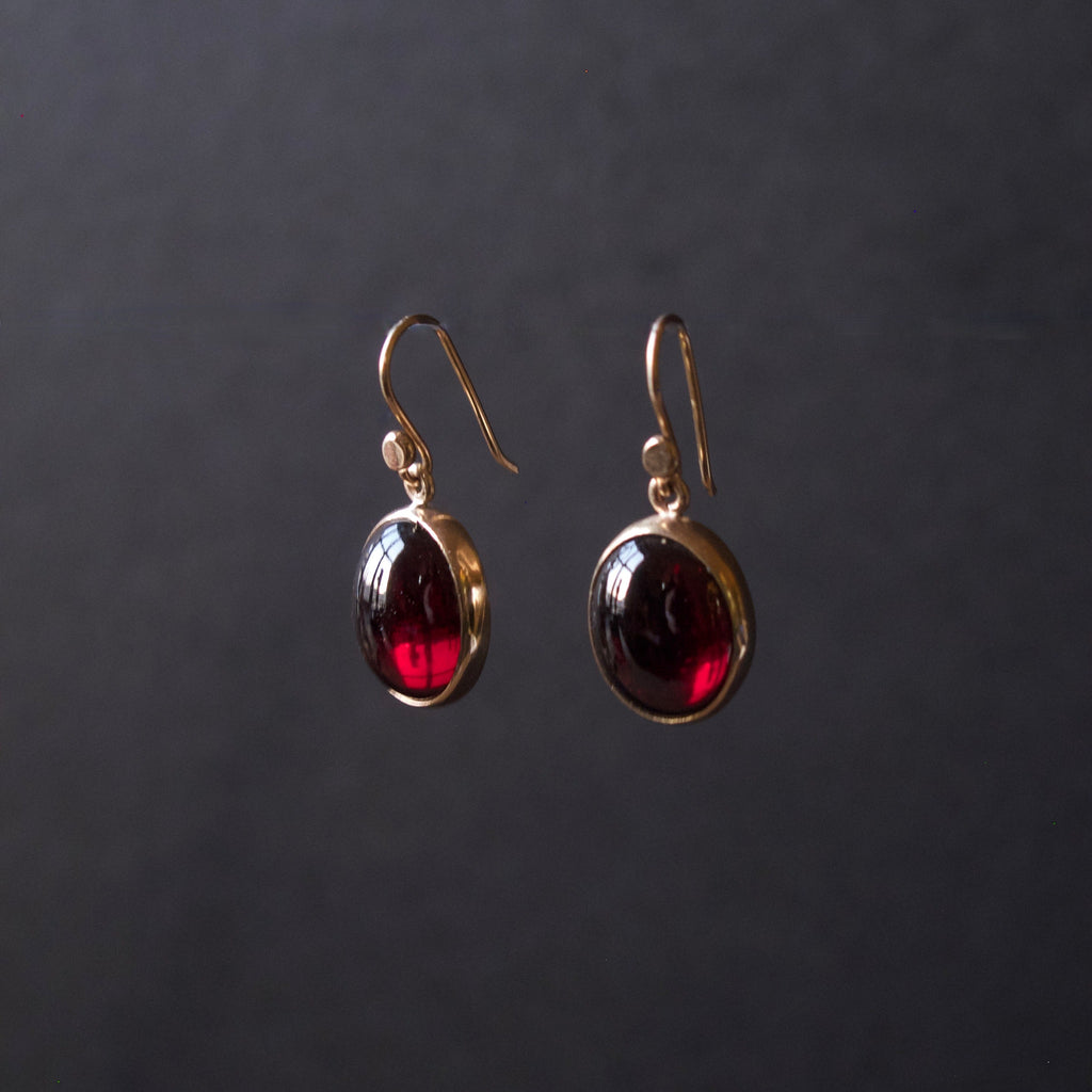 A pair of gold drop earrings with oval dark red gemstones, displayed against a dark gray background.