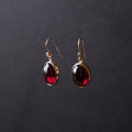 A pair of gold drop earrings with oval dark red gemstones, displayed against a dark gray background.