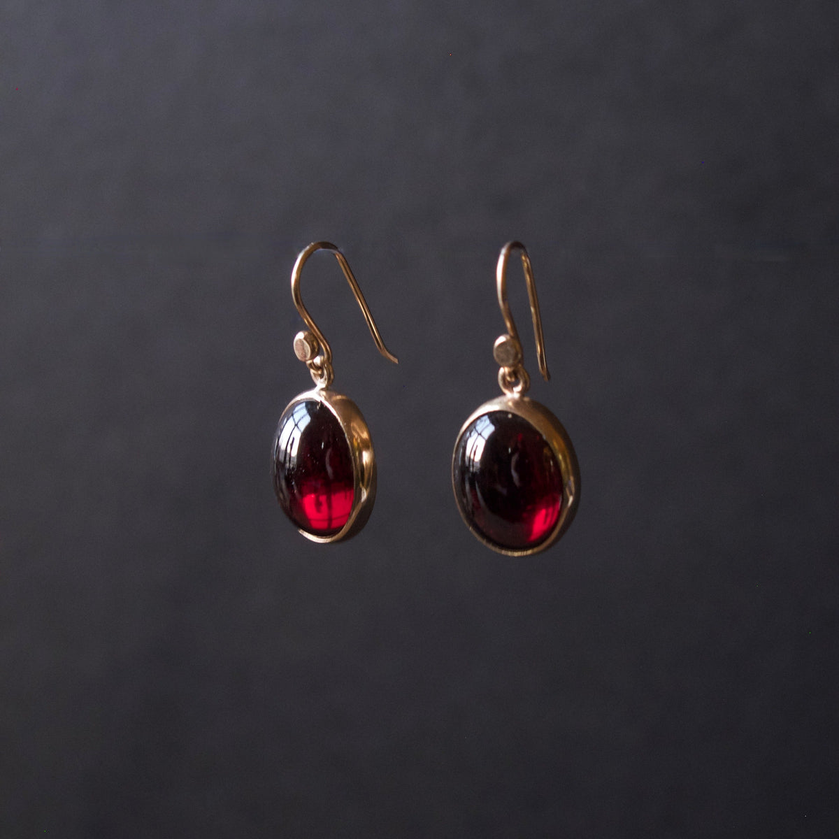 A pair of gold drop earrings with oval dark red gemstones, displayed against a dark gray background.