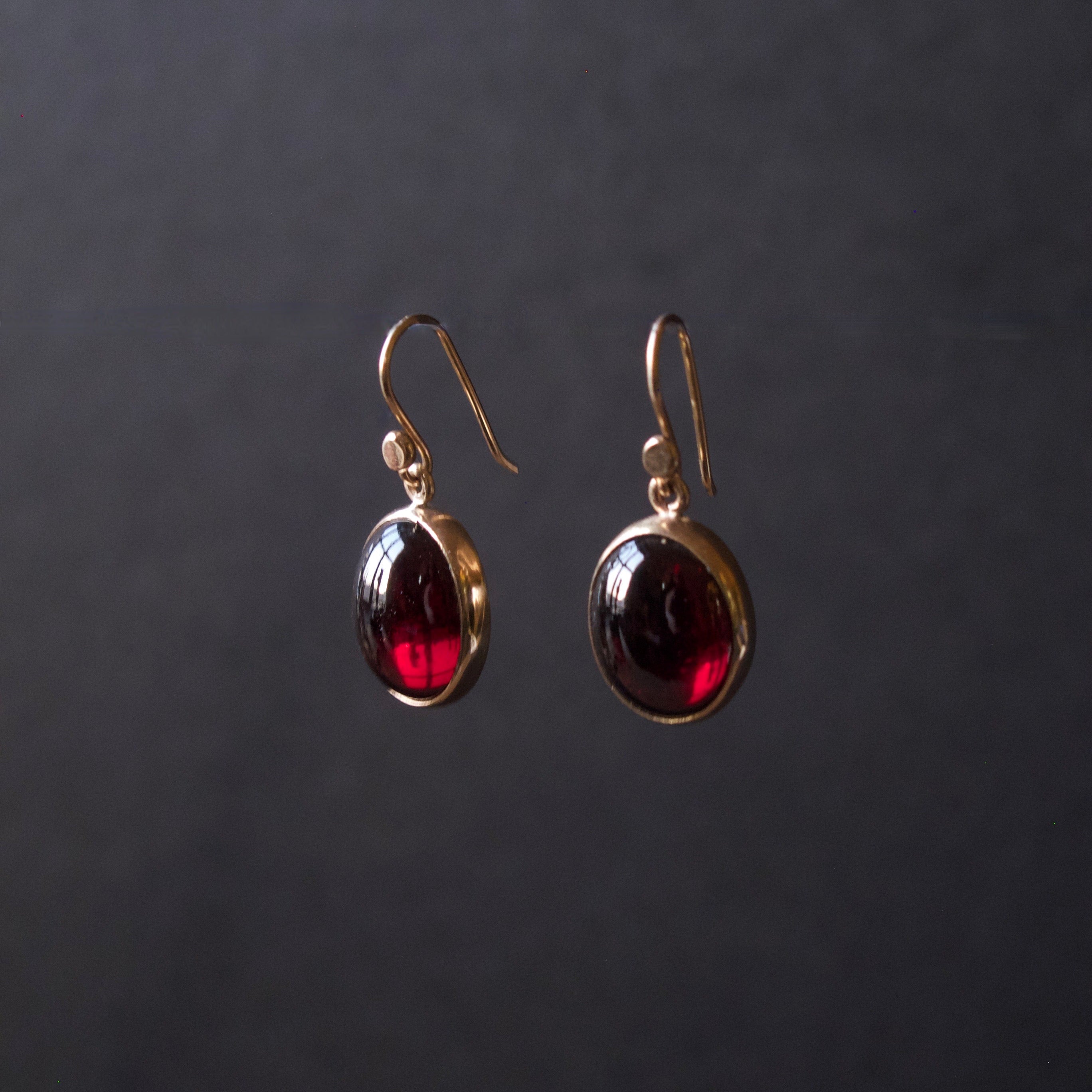 A pair of gold drop earrings with oval dark red gemstones, displayed against a dark gray background.
