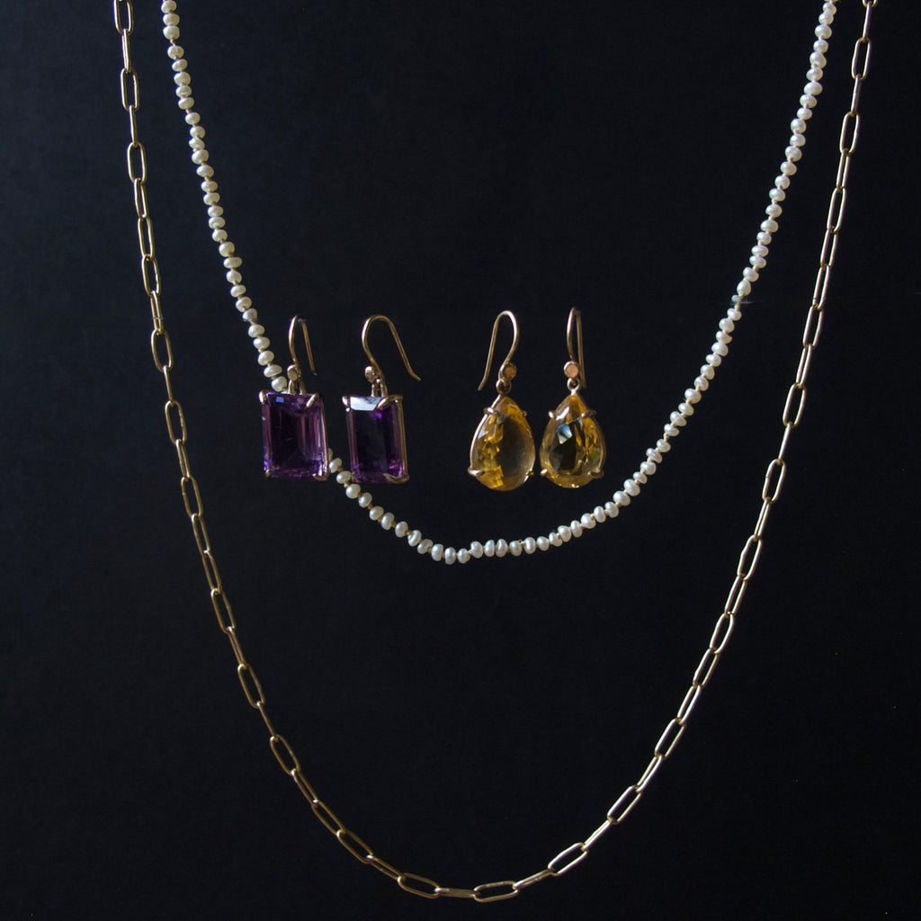 A jewelry set displayed against a black background, featuring a double-strand pearl necklace, a gold paperclip chain, a pair of gold drop earrings with purple rectangular gemstones, and a pair of gold drop earrings with yellow teardrop gemstones.