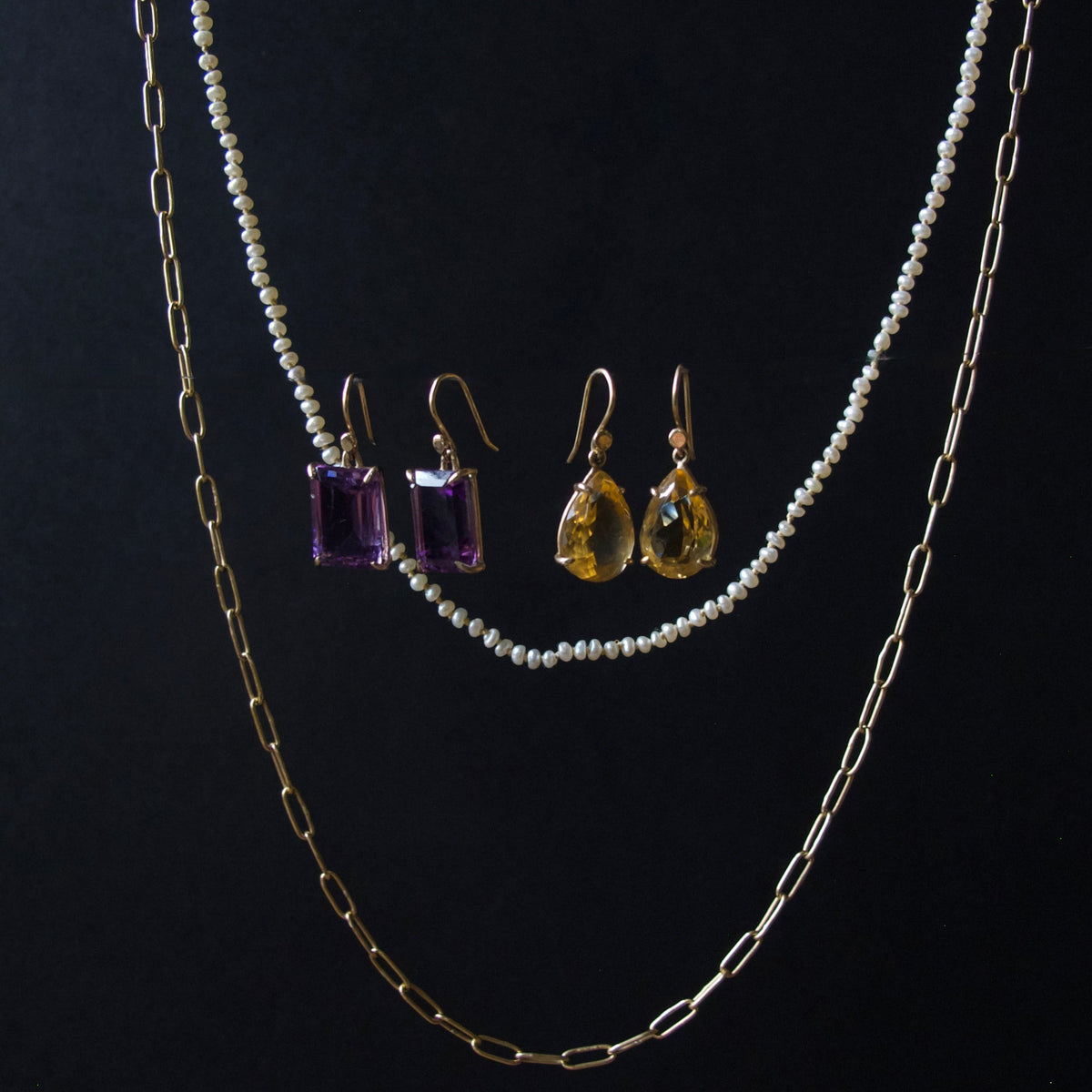 A jewelry set displayed against a black background, featuring a double-strand pearl necklace, a gold paperclip chain, a pair of gold drop earrings with purple rectangular gemstones, and a pair of gold drop earrings with yellow teardrop gemstones.