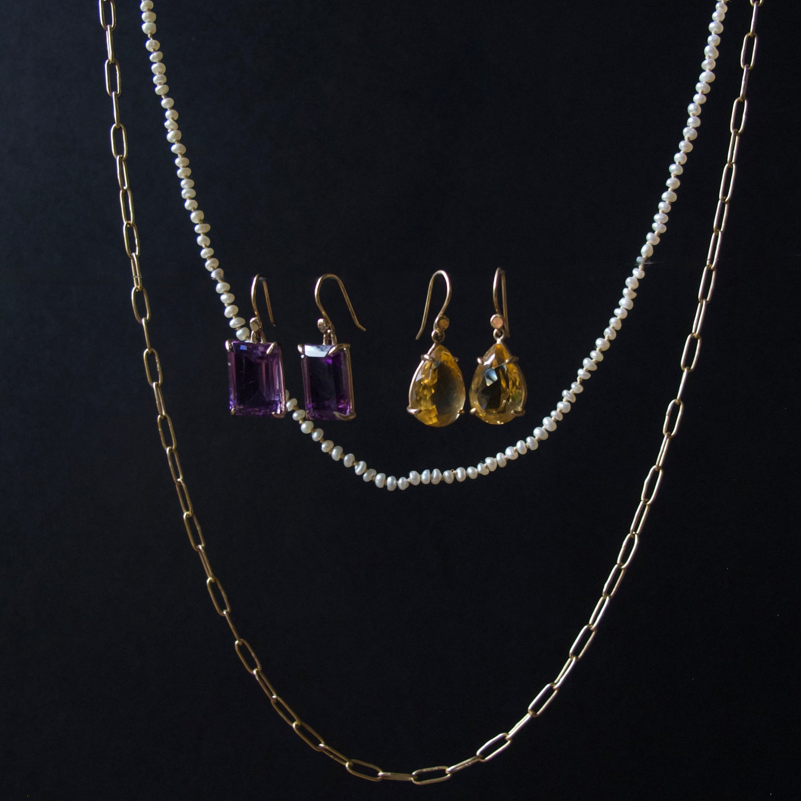 A jewelry set displayed against a black background, featuring a double-strand pearl necklace, a gold paperclip chain, a pair of gold drop earrings with purple rectangular gemstones, and a pair of gold drop earrings with yellow teardrop gemstones.