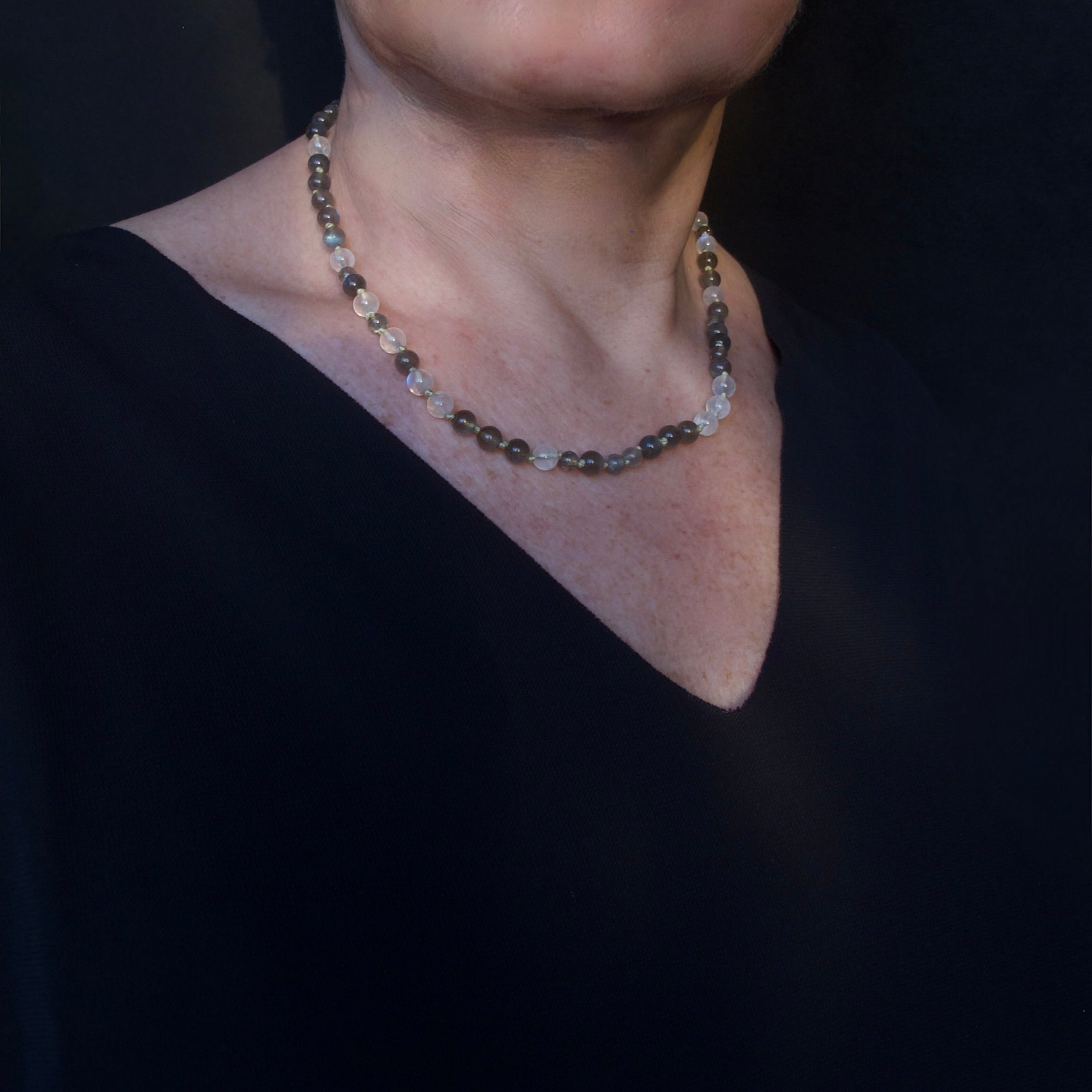 A close-up of a person's neck and lower face wearing a delicate beaded necklace with dark gray, metallic, and clear faceted beads, paired with a black V-neck top against a dark background.