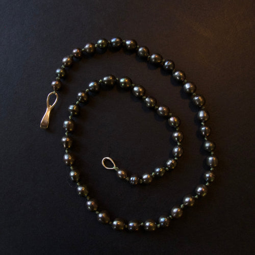 A beaded necklace made of dark, glossy round beads with a small gold-toned clasp, arranged on a dark black background.