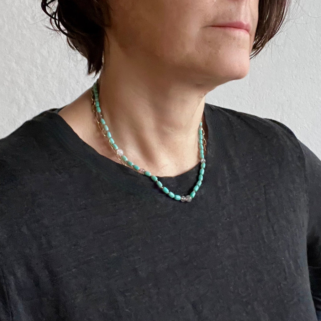 A close-up of a person wearing a double-strand turquoise beaded necklace with small gold and silver spacer beads, paired with a dark charcoal gray t-shirt.