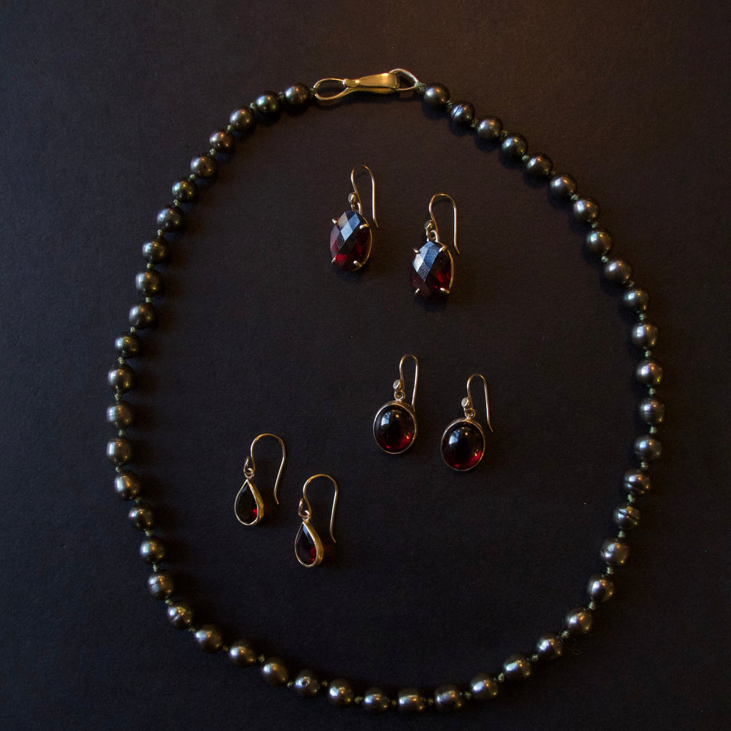 A jewelry set featuring a beaded necklace and four matching drop earrings with dark red teardrop gemstones in gold-toned settings, arranged on a dark background.