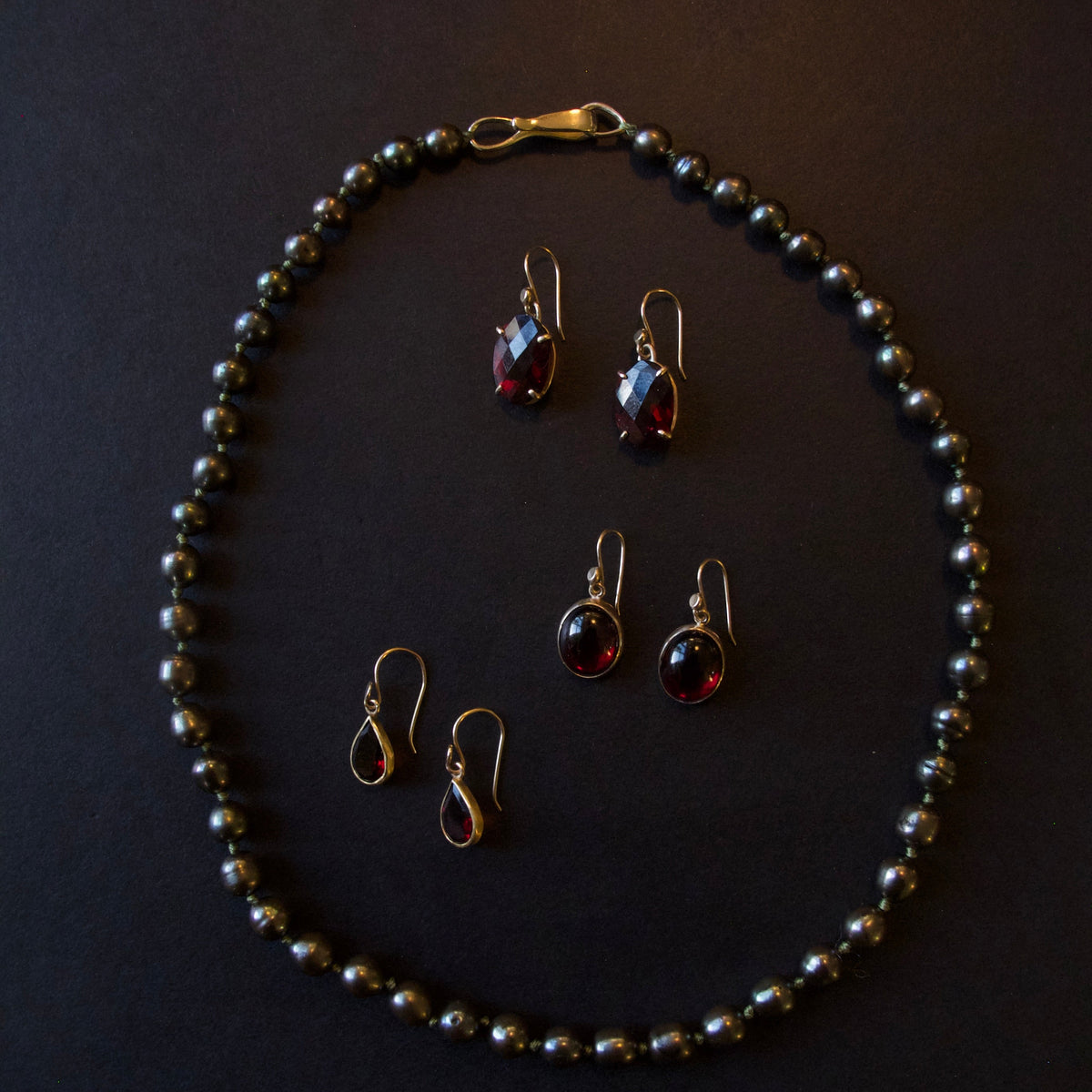 A jewelry set featuring a beaded necklace and four matching drop earrings with dark red teardrop gemstones in gold-toned settings, arranged on a dark background.