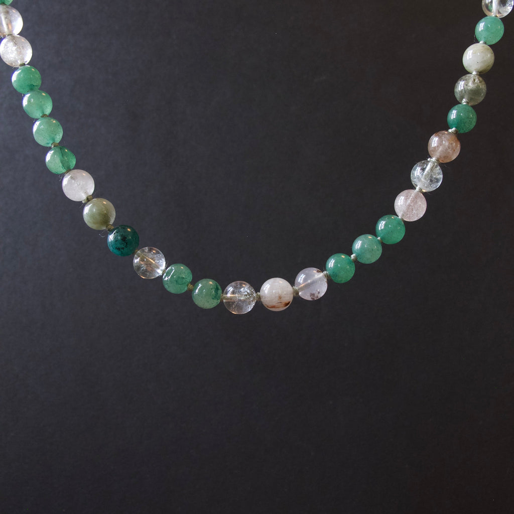A beaded necklace made of round translucent and opaque gemstone beads in shades of green, white, and clear, displayed against a dark black background.