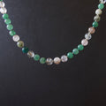 A beaded necklace made of round translucent and opaque gemstone beads in shades of green, white, and clear, displayed against a dark black background.