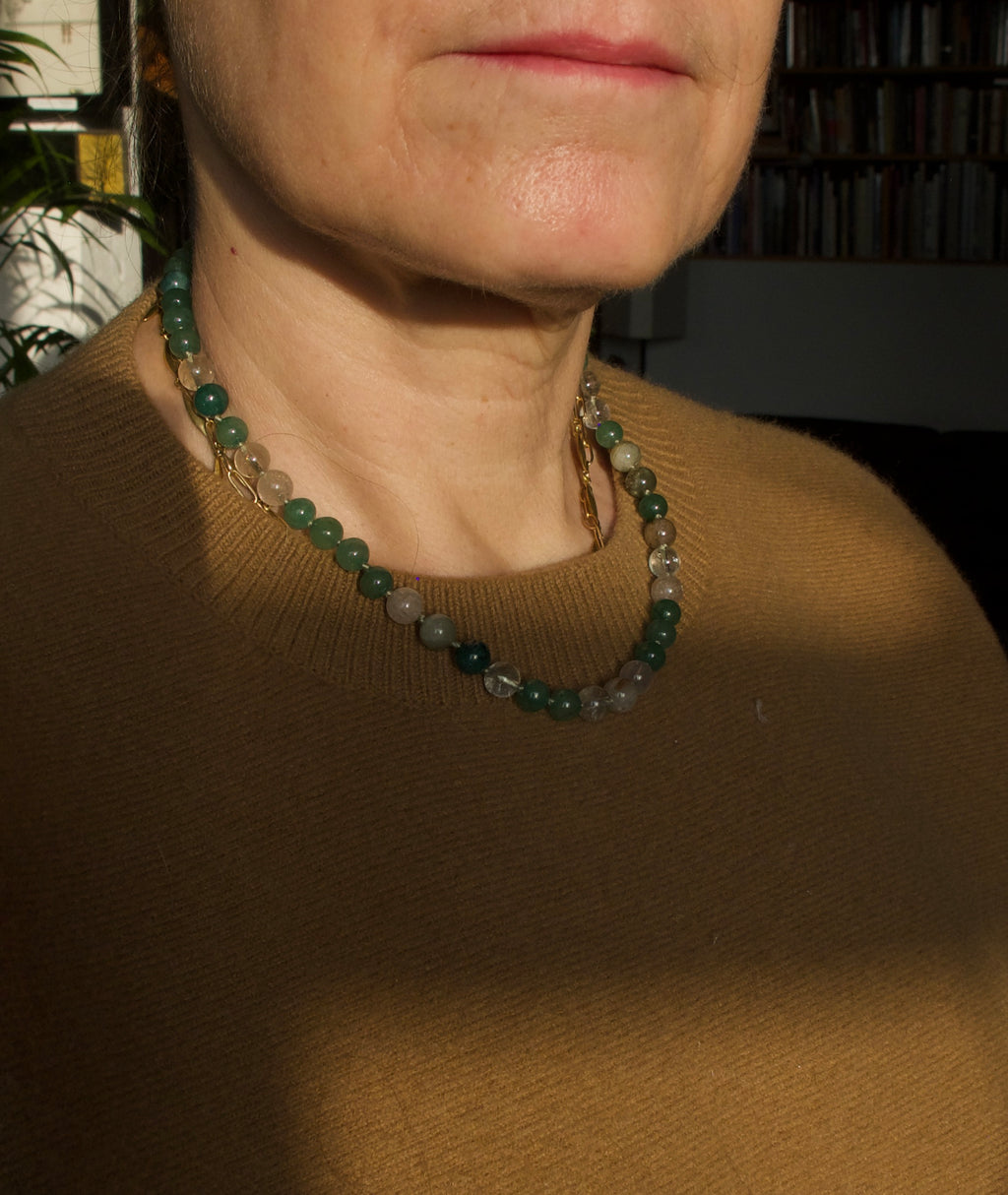 A close-up of a person wearing a beaded necklace made of dark green and translucent gray-green beads, paired with a brown knit sweater. The photo is taken in warm, direct sunlight with a blurred indoor background.