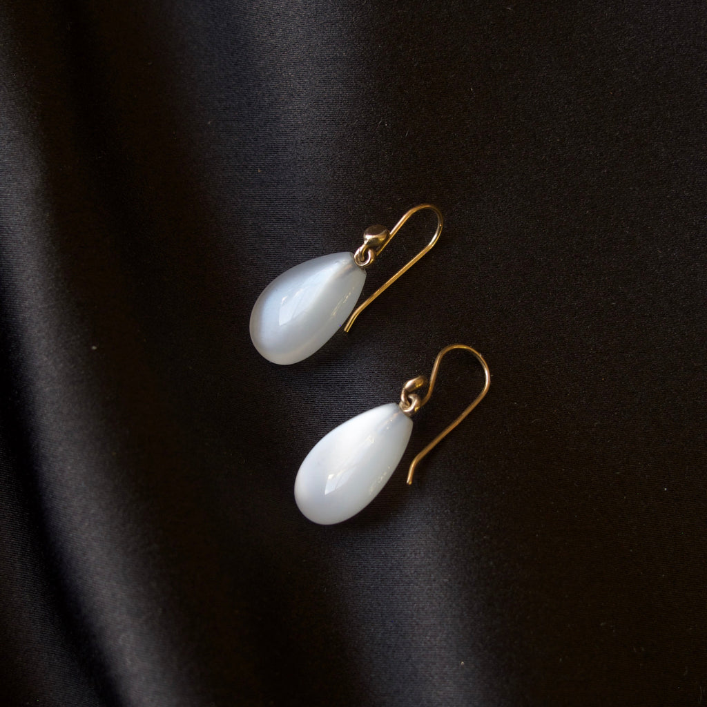 A pair of white teardrop-shaped drop earrings with gold fishhook wires, displayed on a dark black satin fabric background.