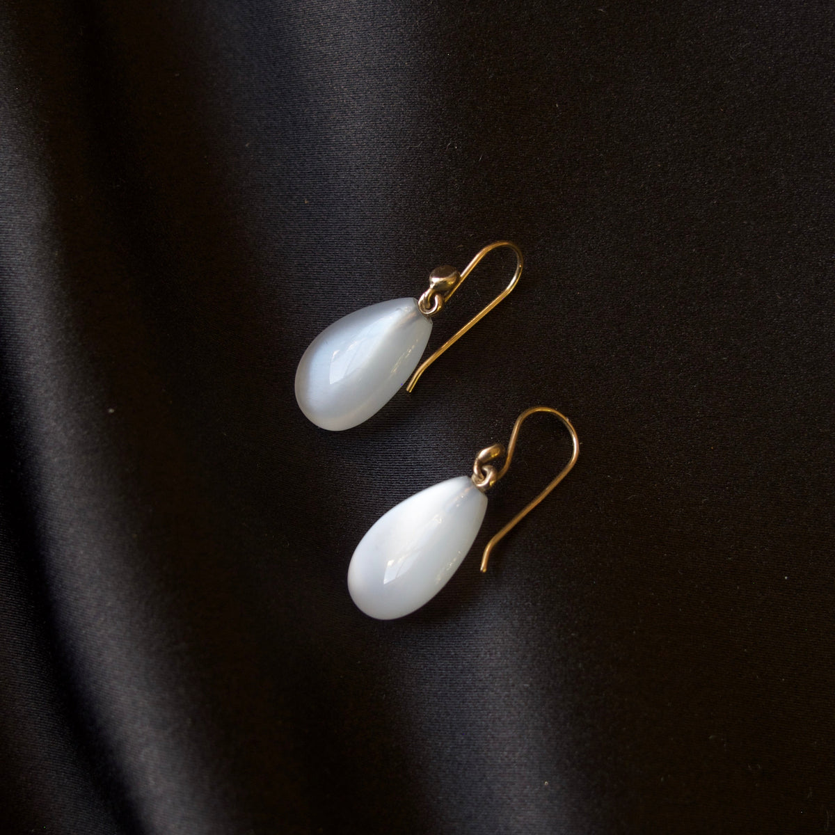 A pair of white teardrop-shaped drop earrings with gold fishhook wires, displayed on a dark black satin fabric background.