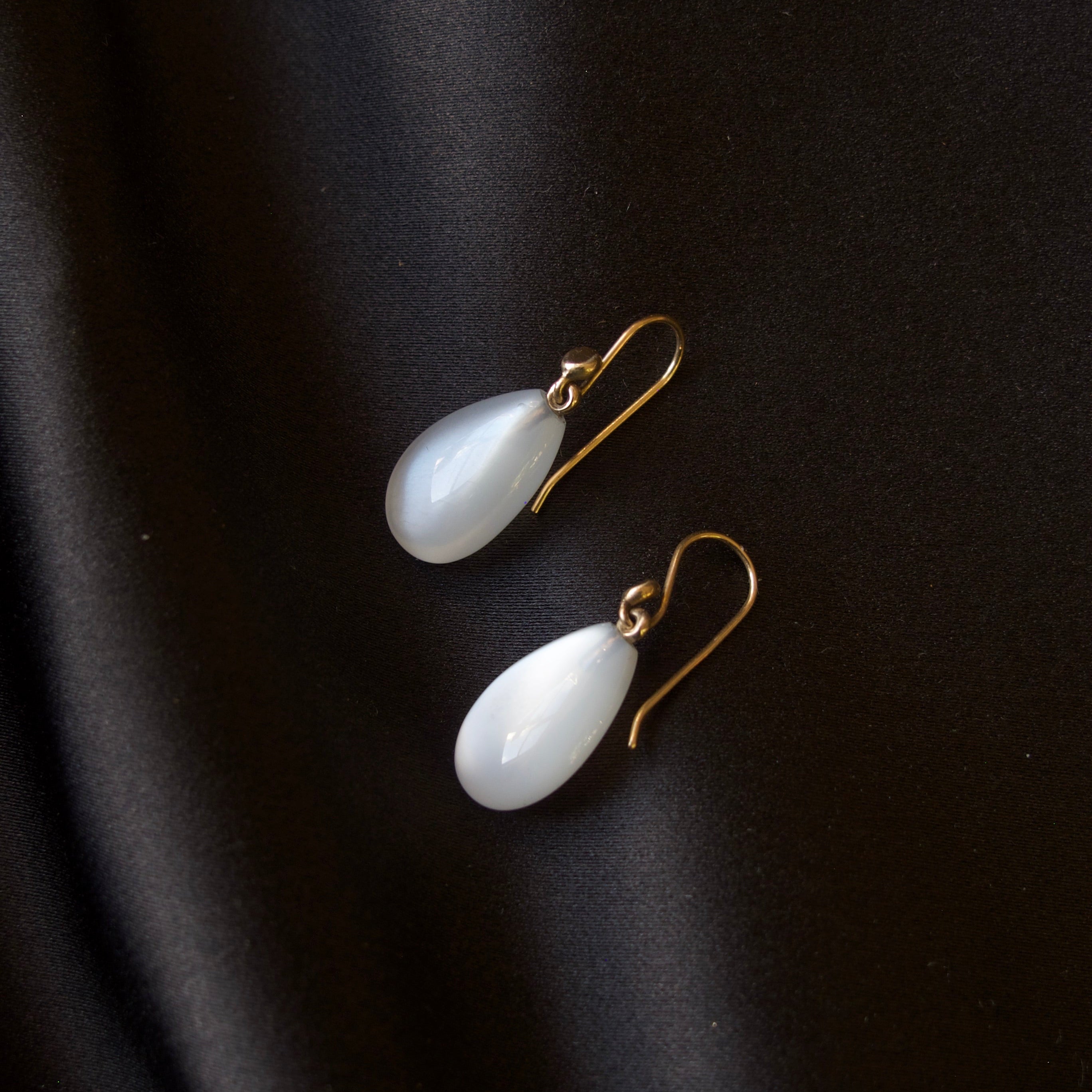 A pair of white teardrop-shaped drop earrings with gold fishhook wires, displayed on a dark black satin fabric background.