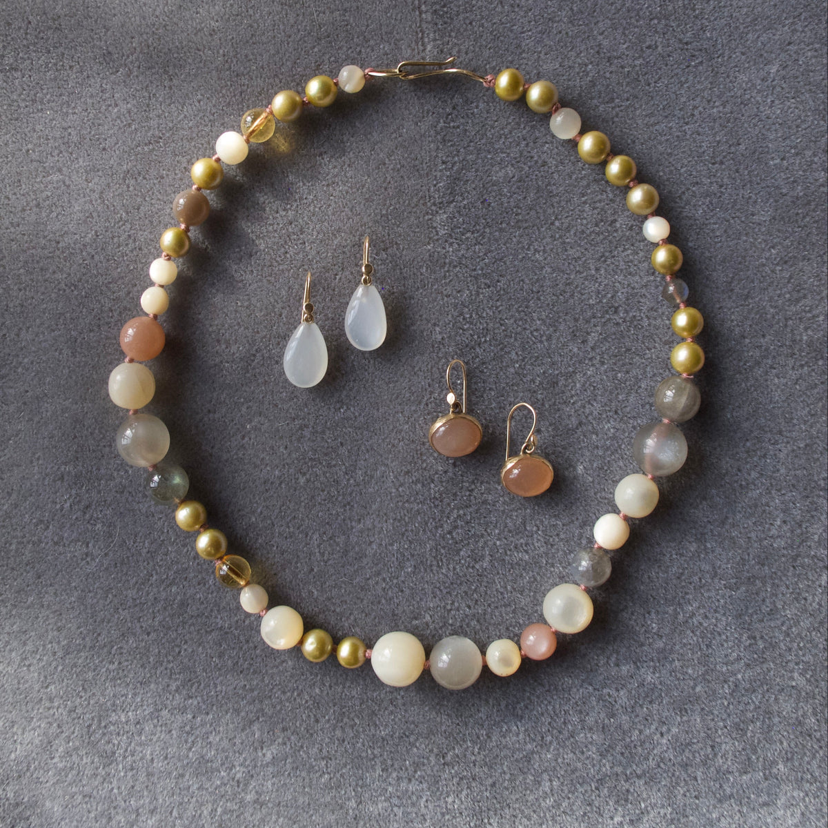 A beaded jewelry set featuring a necklace and matching drop earrings arranged on a textured gray fabric background. The necklace and earrings feature a mix of gold-toned beads, milky white beads, peach-colored beads, and translucent gray beads.