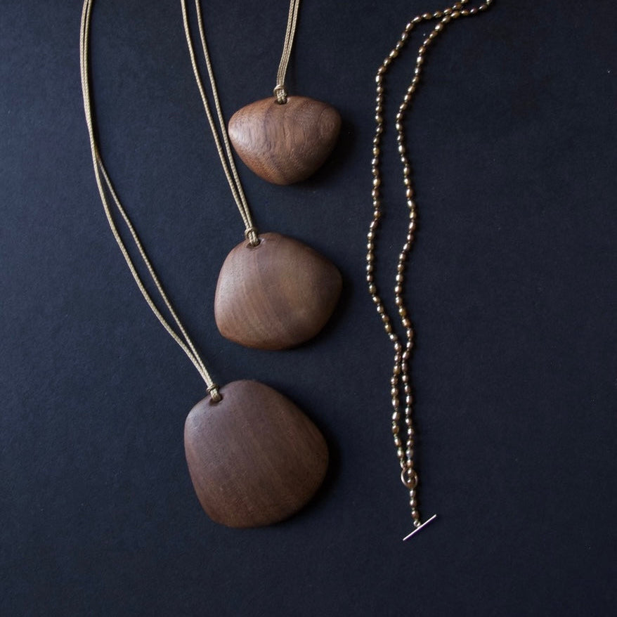 Three wooden pendant necklaces with varying wood grain and sizes, displayed on dark cords against a dark charcoal background.
