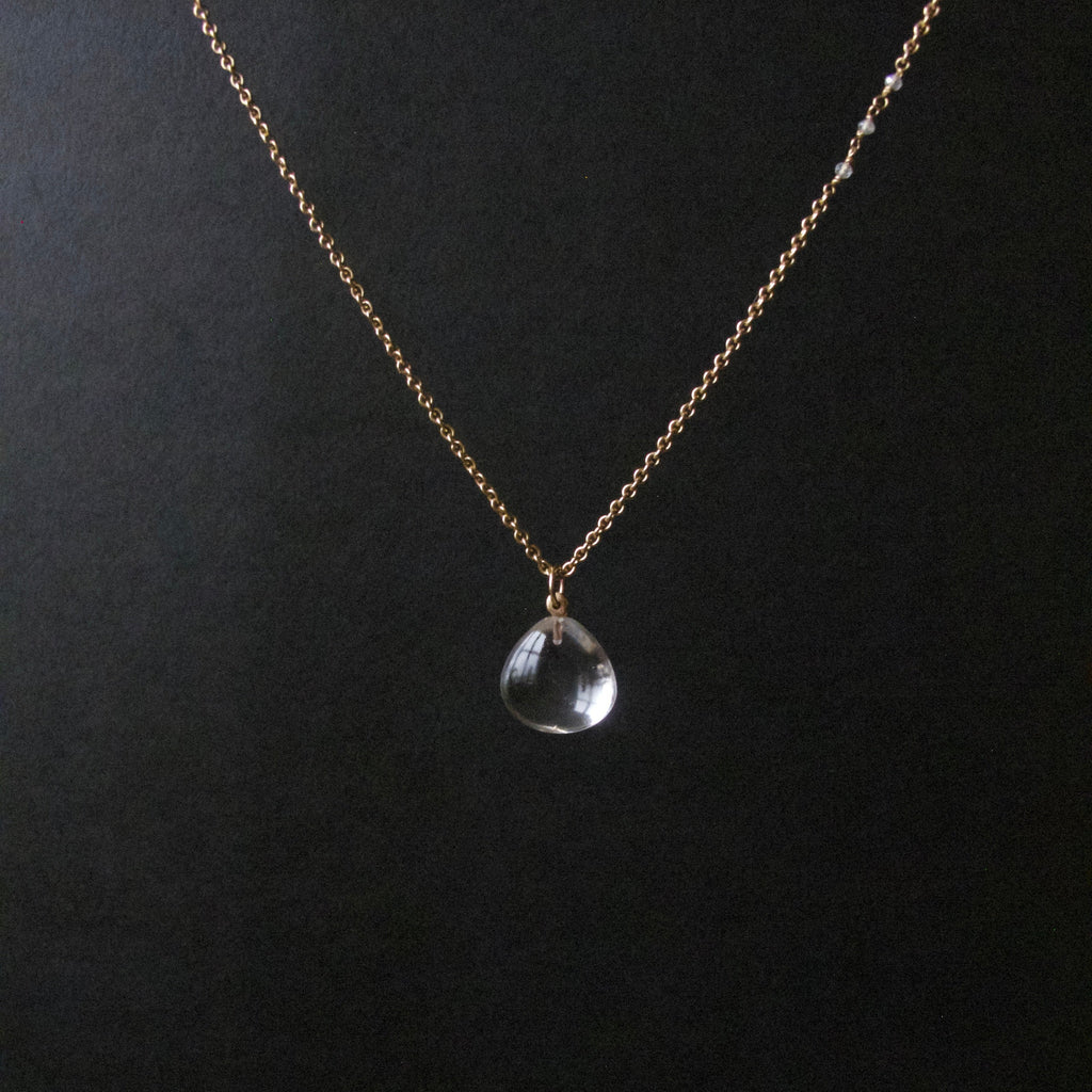 A delicate gold chain necklace with a small clear teardrop-shaped crystal pendant, displayed against a dark black background.