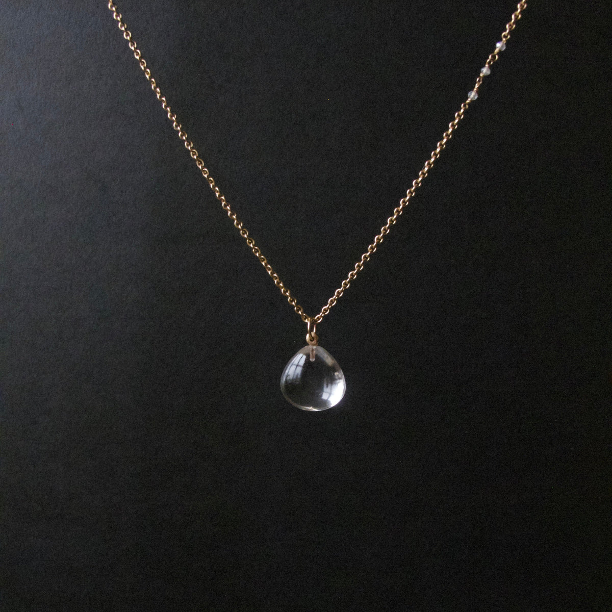 A delicate gold chain necklace with a small clear teardrop-shaped crystal pendant, displayed against a dark black background.