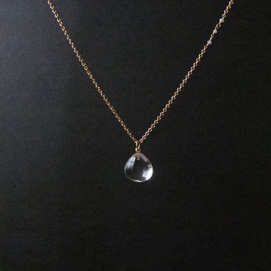 A delicate gold chain necklace with a small clear teardrop-shaped crystal pendant, displayed against a dark black background.
