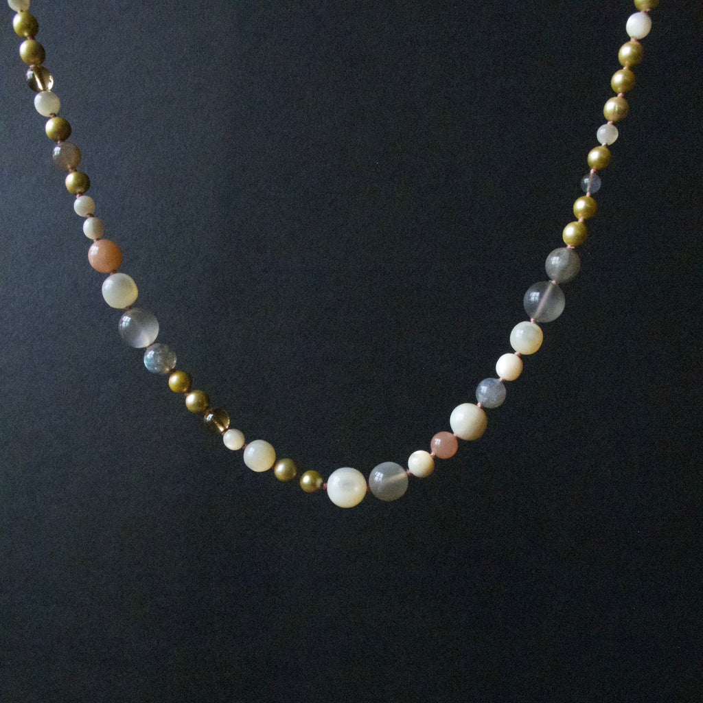 A beaded necklace featuring a mix of matte white, semi-translucent gray, metallic gold, and soft pink beads, displayed against a dark black background.
