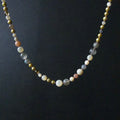 A beaded necklace featuring a mix of matte white, semi-translucent gray, metallic gold, and soft pink beads, displayed against a dark black background.