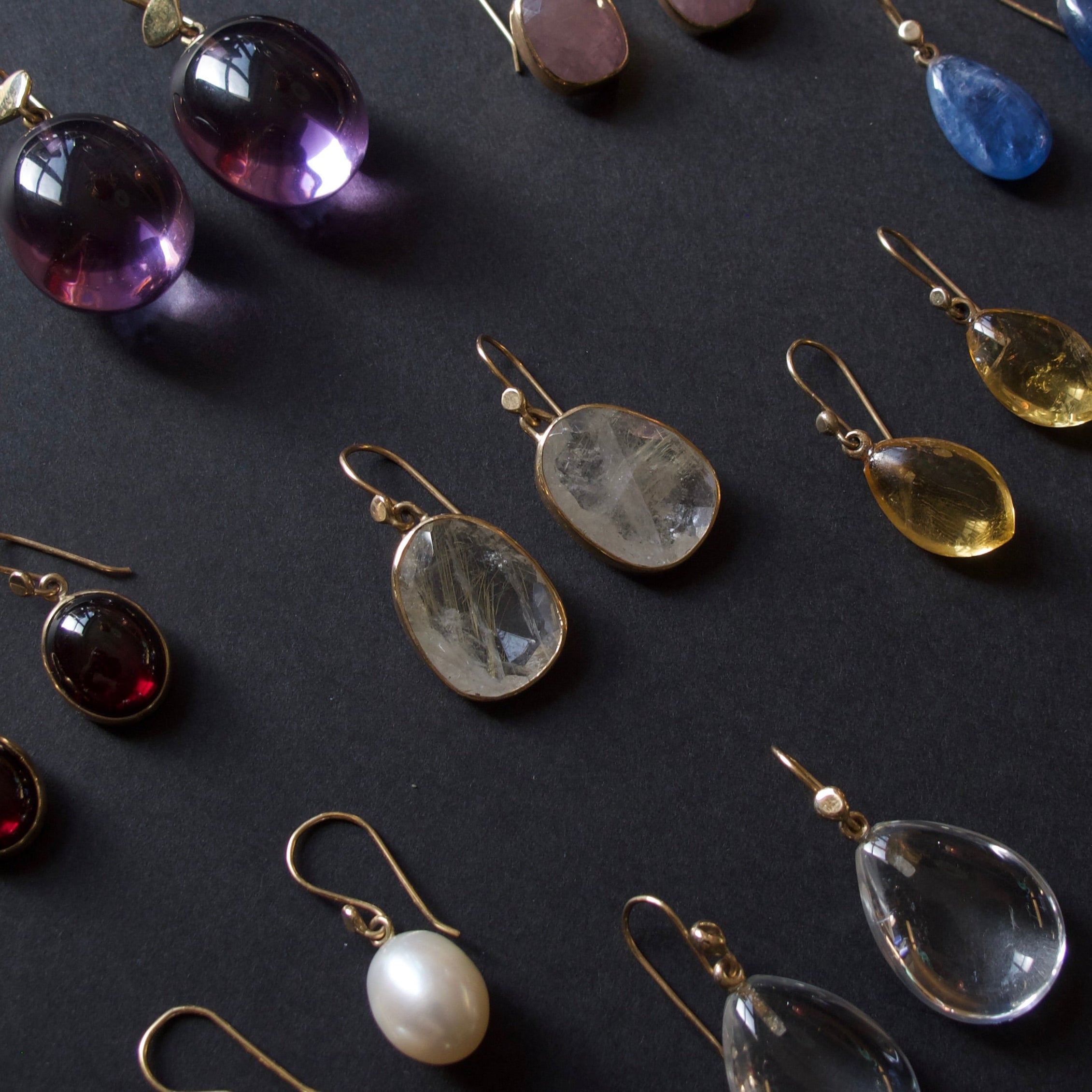 An assortment of colorful gemstone and pearl drop earrings arranged on a dark textured background. The earrings feature various stones including purple amethyst, clear quartz, white pearl, yellow citrine, blue sapphire, and pink rhinestone, set in gold-toned metal.