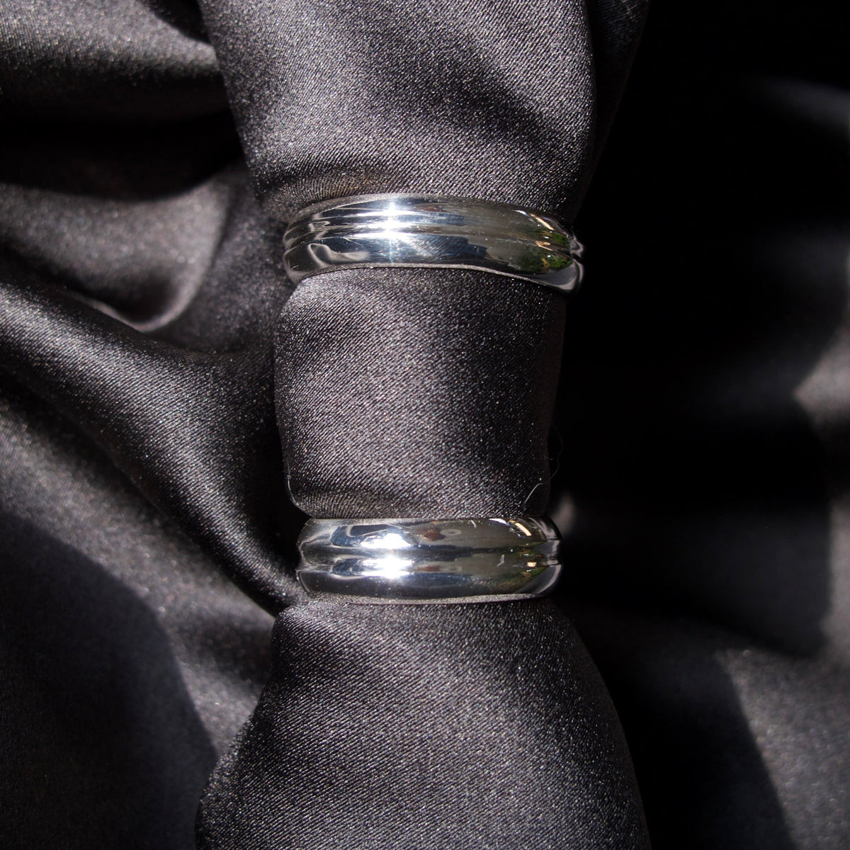 Two polished silver metal bands, likely wedding rings, displayed on a black satin ribbon against a dark background.