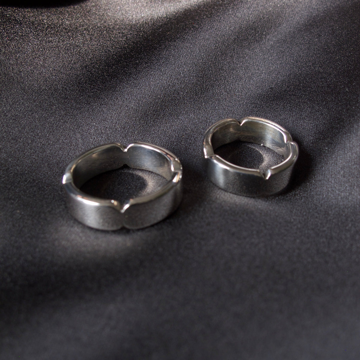 Two silver-toned metal rings with a textured, hammered finish and a small notch detail on the front, displayed on a dark gray satin fabric background.
