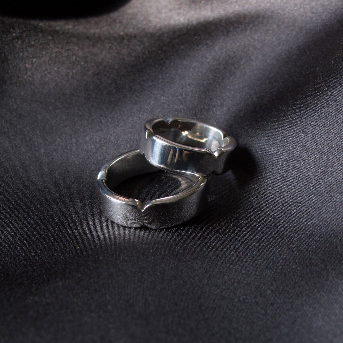 Two silver-toned metal rings with a textured, organic wavy design, resting on a dark, satin-textured surface.