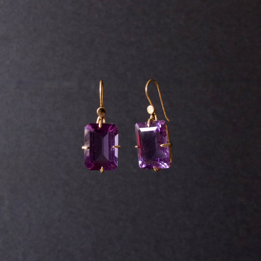 A pair of gold drop earrings featuring rectangular emerald-cut purple gemstones, suspended against a dark gray background.