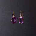 A pair of gold drop earrings featuring rectangular emerald-cut purple gemstones, suspended against a dark gray background.