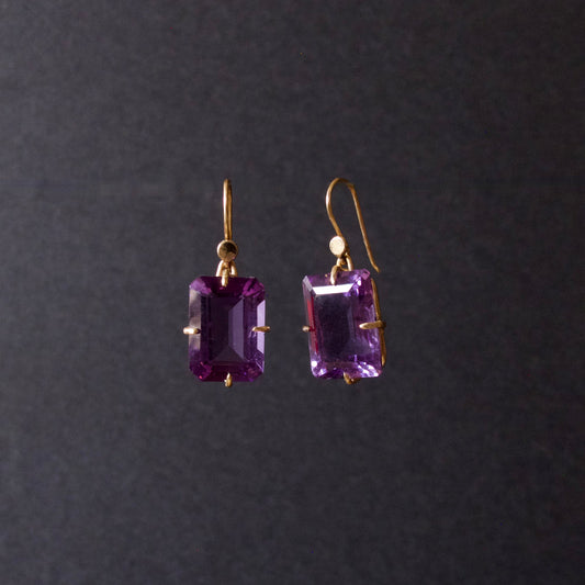 A pair of gold drop earrings featuring rectangular emerald-cut purple gemstones, suspended against a dark gray background.