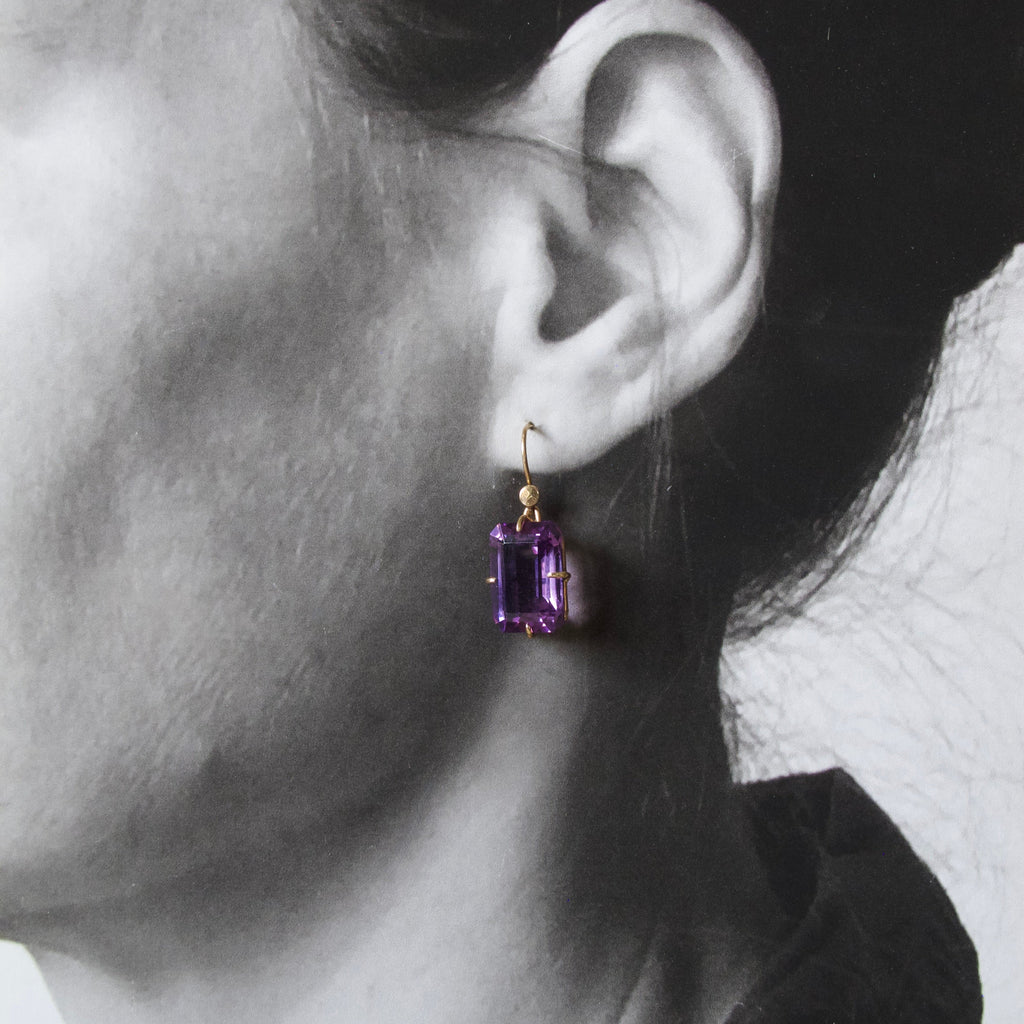 A close-up side profile of a woman wearing a single drop earring with a rectangular purple gemstone in a gold setting.