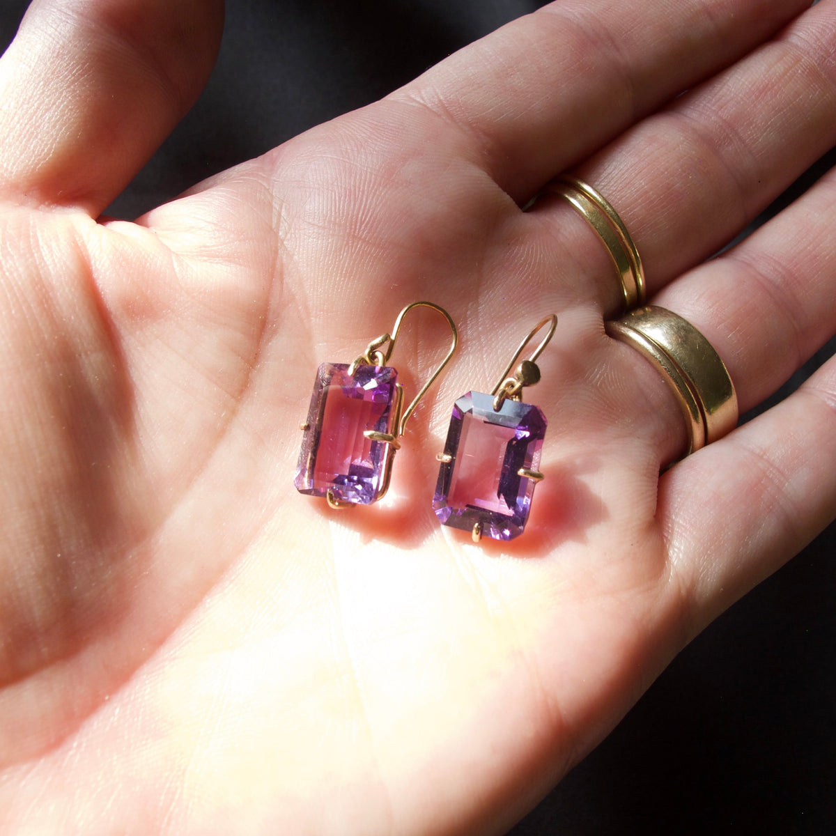A pair of gold drop earrings with rectangular purple gemstones resting on an open hand, with gold rings worn on two fingers against a dark fabric background.