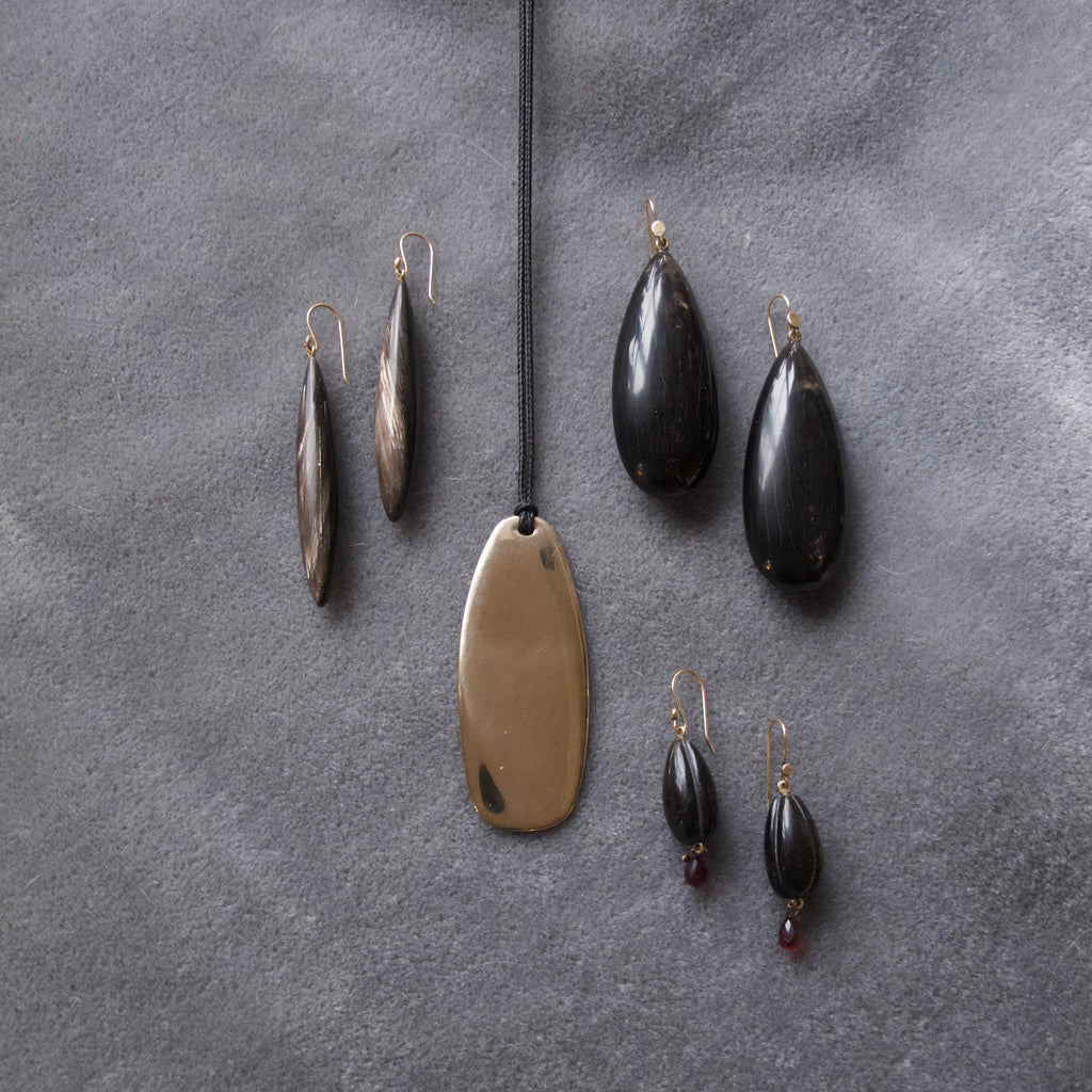 A jewelry set arranged on a textured gray surface, featuring a large oval bronze pendant on a black cord, two dark teardrop earrings, two elongated metallic earrings, and a pair of dark teardrop earrings with small red bead drops.