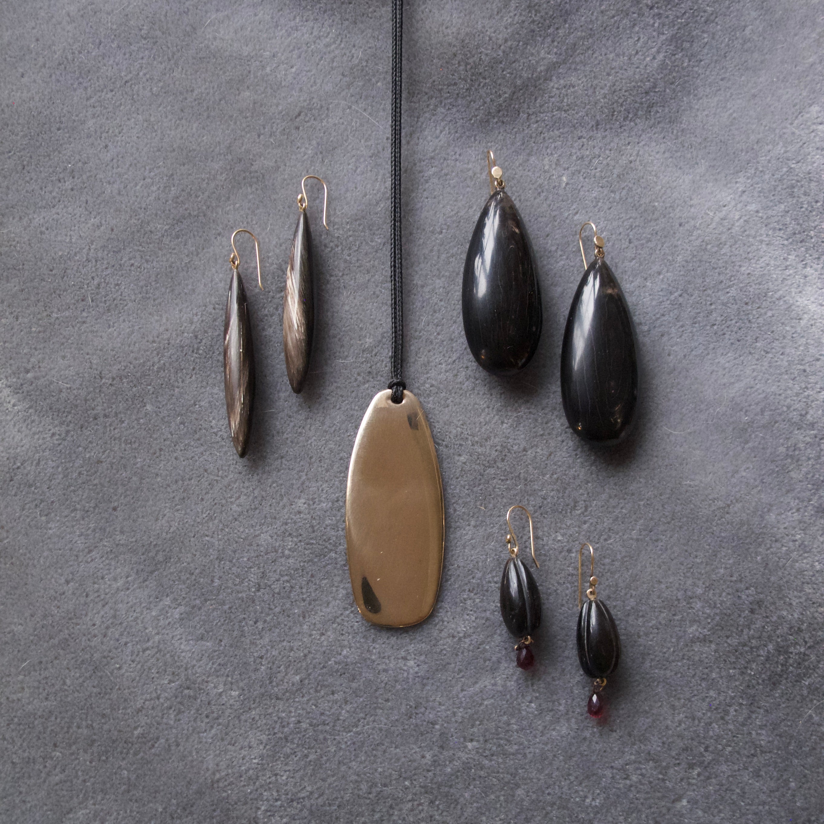 A jewelry set arranged on a textured gray surface, featuring a large oval bronze pendant on a black cord, two dark teardrop earrings, two elongated metallic earrings, and a pair of dark teardrop earrings with small red bead drops.