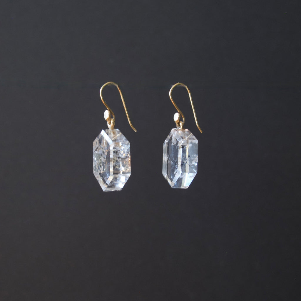 A pair of clear faceted gemstone drop earrings with gold fishhook wires, shown against a solid dark gray background.