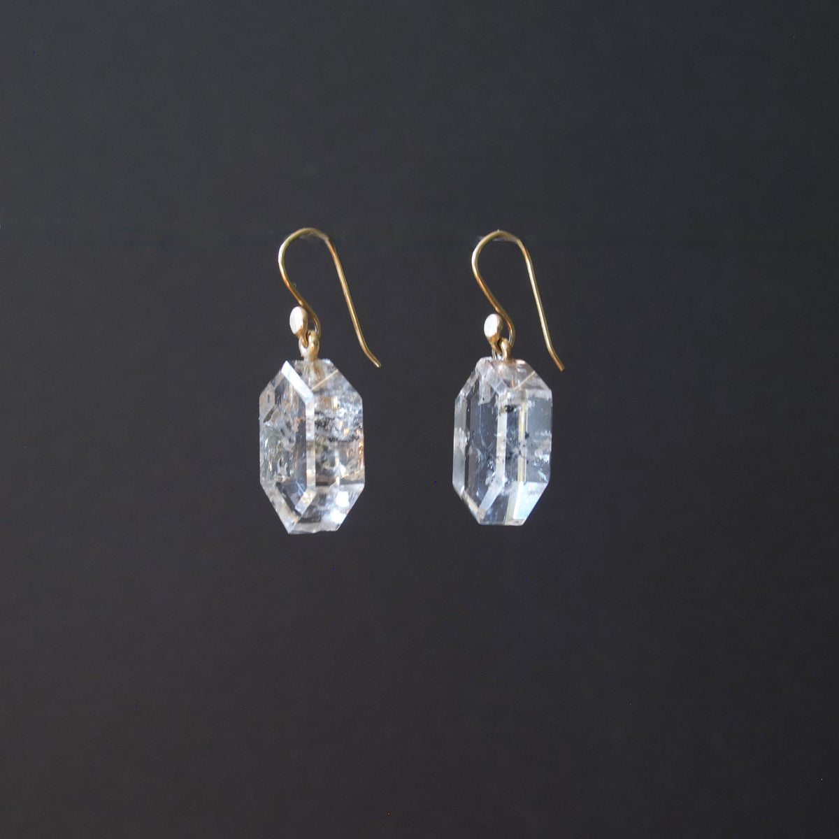 A pair of clear faceted gemstone drop earrings with gold fishhook wires, shown against a solid dark gray background.