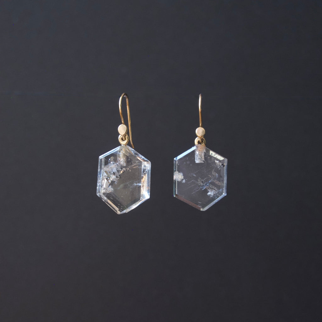 A pair of clear faceted hexagonal gemstone drop earrings with gold fishhook wires and small gold beads, shown against a dark gray background.