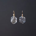 A pair of clear faceted hexagonal gemstone drop earrings with gold fishhook wires and small gold beads, shown against a dark gray background.