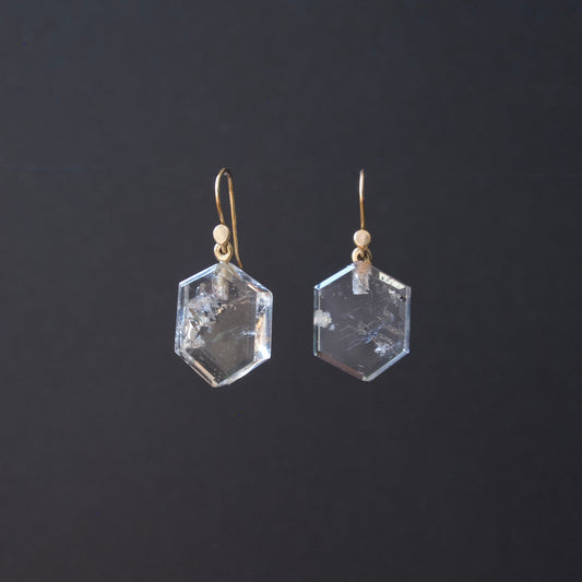 A pair of clear faceted hexagonal gemstone drop earrings with gold fishhook wires and small gold beads, shown against a dark gray background.
