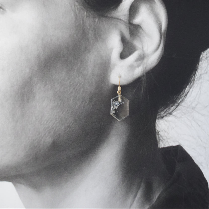 Close-up side profile of a person wearing a small hexagonal clear crystal dangle earring with a gold hook.