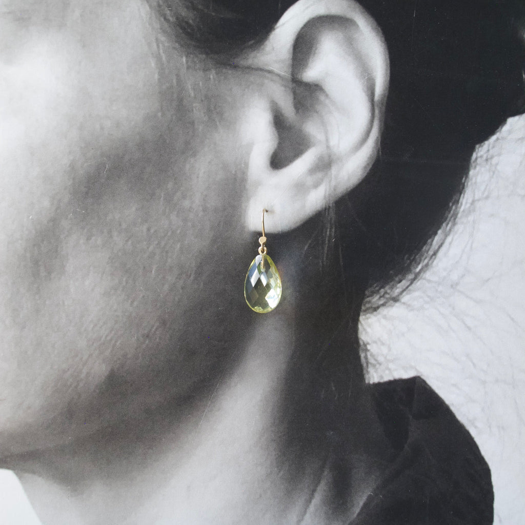 A close-up of a person's ear wearing a small gold teardrop earring with a faceted yellow-green stone.